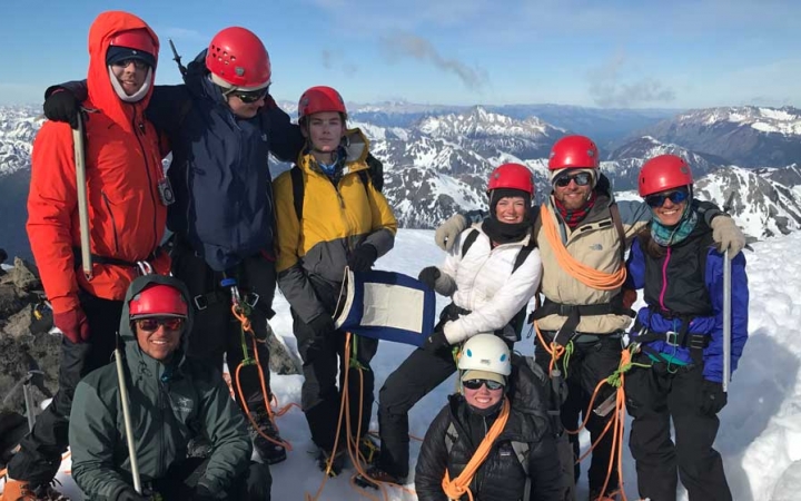 A group of people wearing safety and cold-weather gear stand on a snowy outlook and pose for a photo. Two people are holding a blue peter flag. In the background, there is a snowy, mountainous landscape. 